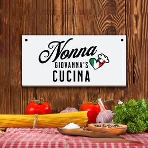 May include: White sign with black text that reads "Nonna Giovanna's Cucina" with a red heart with a green, white, and red Italian flag and a white chef's hat on top. The sign is hanging on a brown wooden wall.