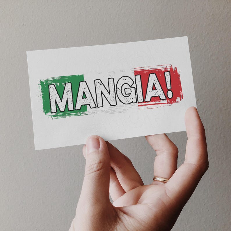 May include: A white card with the Italian phrase "MANGIA!" in bold, outlined letters. The text is set against a green and red brushstroke design, resembling the Italian flag. The card is held by a hand.