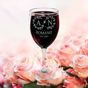 Peut inclure: Un verre à vin transparent rempli de vin rouge, avec les initiales "A" et "N" dans une couronne florale. Le nom "Romano" et l'année "EST. 2025" sont également affichés. Le verre est posé sur un fond de roses roses.