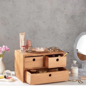 Puede incluir: Un organizador de maquillaje de madera con cuatro cajones, algunos abiertos, que muestran cosméticos. El organizador es de madera clara y se encuentra sobre una superficie blanca. Varios productos de belleza, incluyendo esmalte de uñas y un espejo, están dispuestos alrededor.
