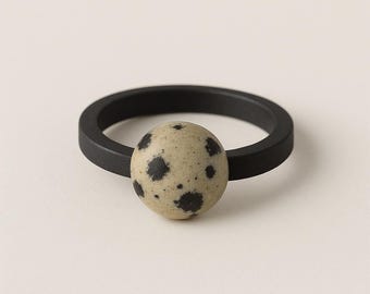 May include: A black ring with a speckled, light-colored sphere at the center. The sphere has dark spots, and the ring band is a solid, matte black. The ring is set against a neutral background.