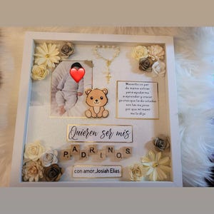 May include: A white picture frame with a gold border, decorated with paper flowers and a small teddy bear. The frame contains a photo of a baby, a rosary, and a sign that reads "Quieren ser mis PADRINOS con amor, Josiah Elias".