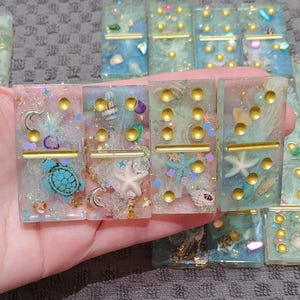 May include: A set of handmade resin dominoes with a beach theme. The dominoes are translucent and feature gold glitter, seashells, starfish, and a turtle.