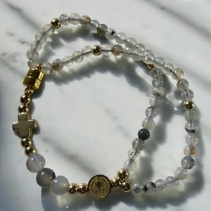 May include: A rosary necklace featuring translucent, speckled beads and gold-colored accents. The necklace includes a gold cross pendant and a medallion. The beads are strung on a flexible cord, creating a loop.