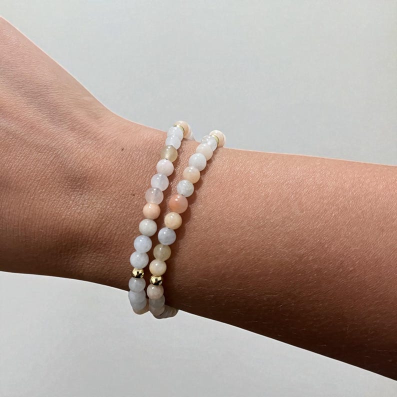 May include: Two beaded bracelets with a mix of pastel-colored beads, including white, peach, and pale blue. The bracelets are strung on elastic and have small gold-colored accents. The bracelets are worn on a wrist.