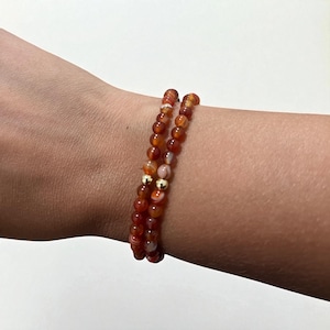 May include: Two beaded bracelets with orange and white stones and gold-colored accents. The bracelets are worn on a wrist. The beads are small and round, strung together to form a flexible band. The bracelets are a warm, earthy color.