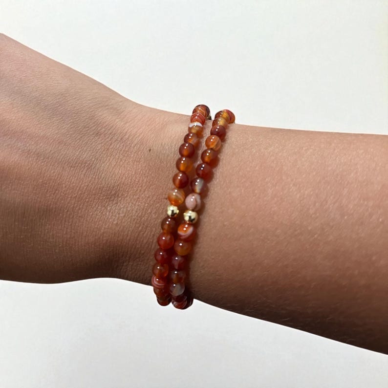 May include: Two beaded bracelets with orange and white stones and gold-colored accents. The bracelets are worn on a wrist. The beads are small and round, strung together to form a flexible band. The bracelets are a warm, earthy color.