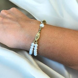 May include: Gold-coloured rosary bracelet with white beads and a gold cross charm. The bracelet includes a gold centrepiece with a religious figure. The design is simple and elegant, suitable for religious occasions or everyday wear.