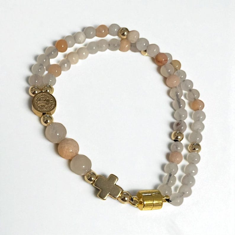 May include: A double-strand bracelet featuring gray and peach-colored beads. The bracelet includes gold-colored accents, a cross charm, and a magnetic clasp. The beads are strung on a flexible cord.
