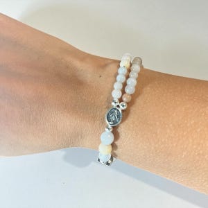 May include: A beaded bracelet with white, light pink, and pale yellow beads. The bracelet features a silver-toned centerpiece with a religious emblem and a silver-toned square charm.