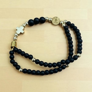 Könnte beinhalten: Schwarzes Perlenarmband mit einem goldfarbenen Kreuz und einem goldfarbenen Medaillon. Das Armband besteht aus mattschwarzen Perlen und kleinen, glänzenden goldfarbenen Perlen. Das Armband liegt auf einer hellen Holzoberfläche.