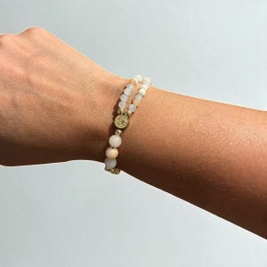 May include: A double-strand bracelet featuring light pink and white beads. The bracelet is accented with gold-colored metal spacers and a central decorative element. The beads appear to be natural stone, and the bracelet is worn on a wrist.