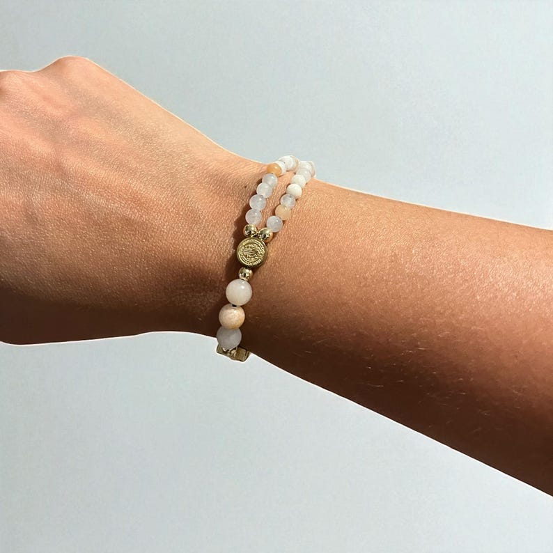 May include: A double-strand bracelet featuring light pink and white beads. The bracelet is accented with gold-colored metal spacers and a central decorative element. The beads appear to be natural stone, and the bracelet is worn on a wrist.