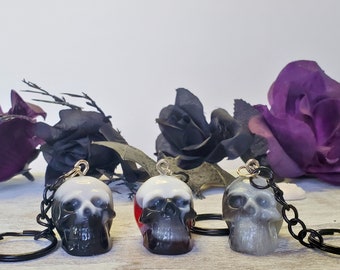 May include: Three skull-shaped keychains with black chains and key rings. The skulls are white, gray, and black, with a gradient effect. The background features black and purple roses.
