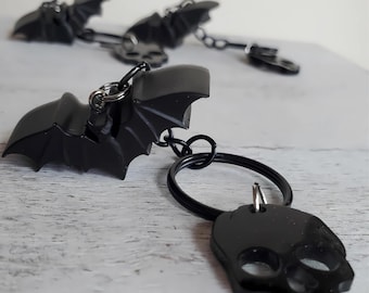 Pode incluir: Chaveiros pretos de morcego e caveira. Os chaveiros de morcego têm asas detalhadas e são presos a uma corrente. O chaveiro de caveira tem o formato de uma caveira detalhada. Os chaveiros são presos a um chaveiro preto.