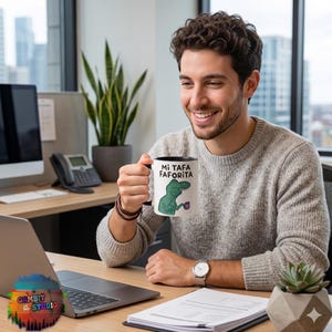 May include: White ceramic mug with a black interior and handle. The mug features the text "MI TAFA FAVORITA" and a cartoon dinosaur holding a cup. The person in the image is smiling and holding the mug.