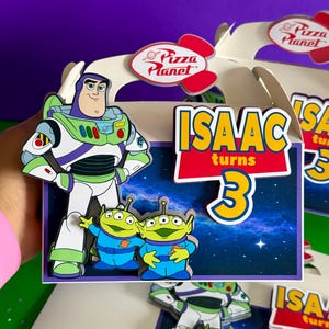 May include: A birthday party decoration featuring Buzz Lightyear and the Little Green Men from Toy Story. The box has the text "ISAAC turns 3" in yellow and blue. The Pizza Planet logo is also visible.