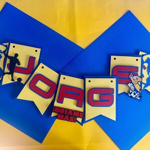 May include: A yellow and red banner with the word "JOGA" in red letters. The banner is decorated with soccer-themed images, including a soccer ball, a player, and a cartoon eagle. The banner also includes the text "ODIAME MAS!" in red letters.