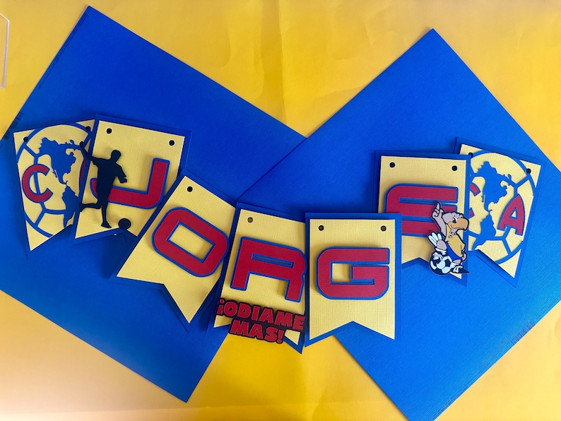 May include: A yellow and red banner with the word "JOGA" in red letters. The banner is decorated with soccer-themed images, including a soccer ball, a player, and a cartoon eagle. The banner also includes the text "ODIAME MAS!" in red letters.
