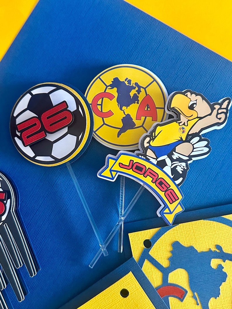 K&ouml;nnte beinhalten: Drei Cupcake-Topper mit einem Fu&szlig;ball mit der Nummer 26, einem gelben und blauen Fu&szlig;ball-Teamlogo mit den Buchstaben CA und einem gelben Cartoon-Vogel, der ein blaues und gelbes Fu&szlig;balltrikot mit dem Namen Jorge auf einem Banner tr&auml;gt.