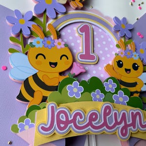 May include: A colorful cake topper featuring cartoon bees, flowers, and the name "Jocelyn." The design includes a number "1" and a rainbow arch, perfect for a first birthday celebration. The bees are yellow and black, with blue wings and flower crowns.