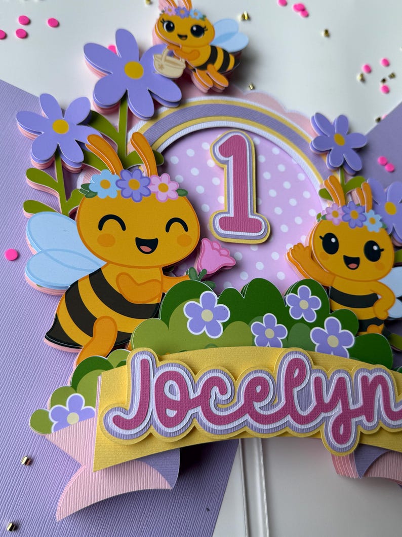 May include: A colorful cake topper featuring cartoon bees, flowers, and the name "Jocelyn." The design includes a number "1" and a rainbow arch, perfect for a first birthday celebration. The bees are yellow and black, with blue wings and flower crowns.