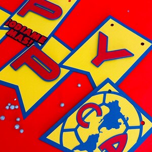 May include: A yellow and blue banner with the word "Happy" in red letters. The banner also includes a red and blue soccer ball with the letters "CA" in red. The text "&iexcl;ODIAME MAS!" is also on the banner.