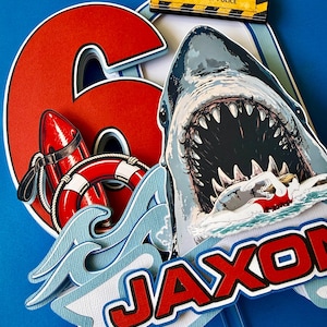 May include: A layered cake topper featuring a large red number 6, a shark with an open mouth, a red life buoy, and a swimmer. It includes a yellow sign that reads BEACH CLOSED BY ORDER OF POLICE and a banner with the name JAXON in red letters.