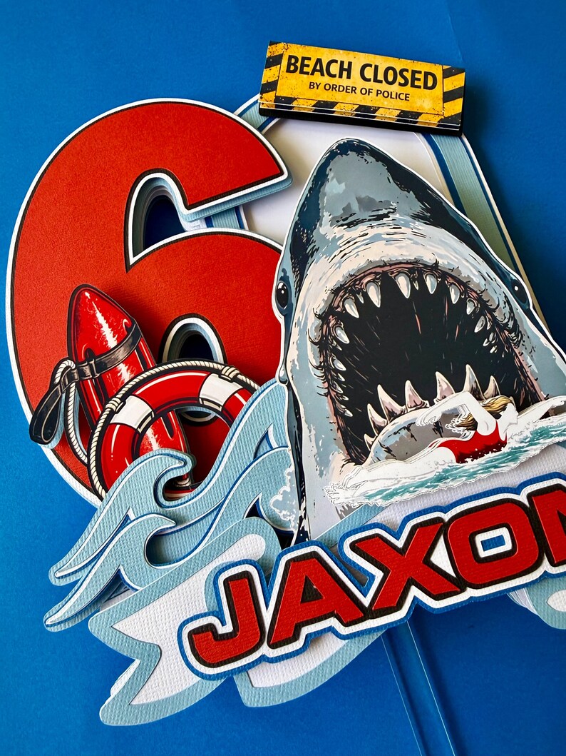 May include: A layered cake topper featuring a large red number 6, a shark with an open mouth, a red life buoy, and a swimmer. It includes a yellow sign that reads BEACH CLOSED BY ORDER OF POLICE and a banner with the name JAXON in red letters.