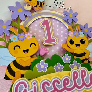 May include: A colorful birthday cake topper featuring a bee theme. The design includes smiling bees, flowers, the number "1", and the name "Giselle" in pink and white lettering. The color palette includes yellow, pink, purple, and green.