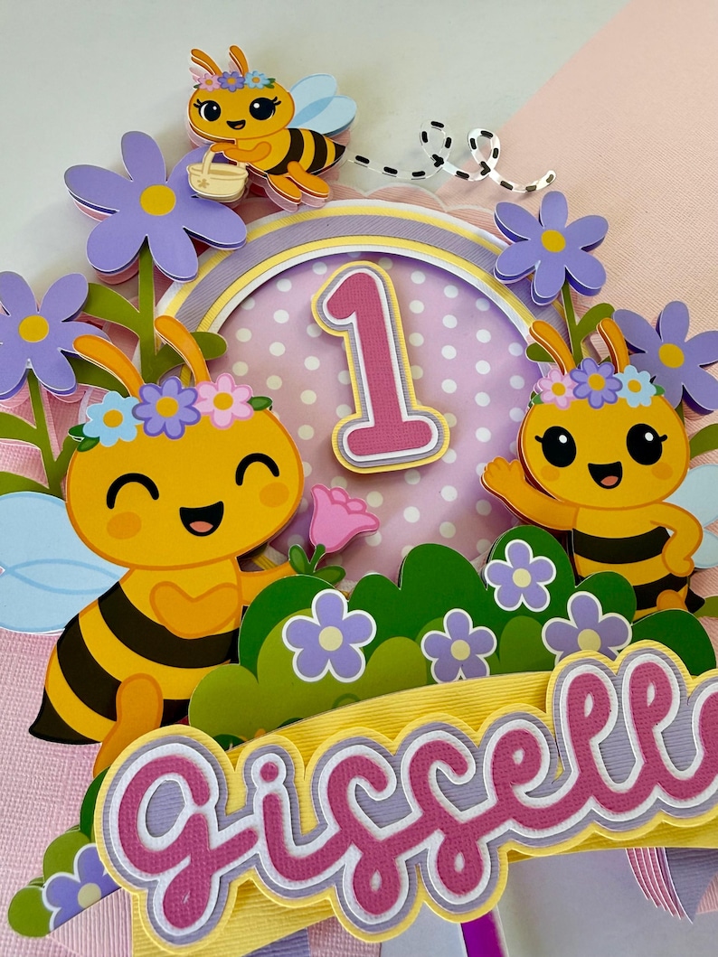 May include: A colorful birthday cake topper featuring a bee theme. The design includes smiling bees, flowers, the number "1", and the name "Giselle" in pink and white lettering. The color palette includes yellow, pink, purple, and green.