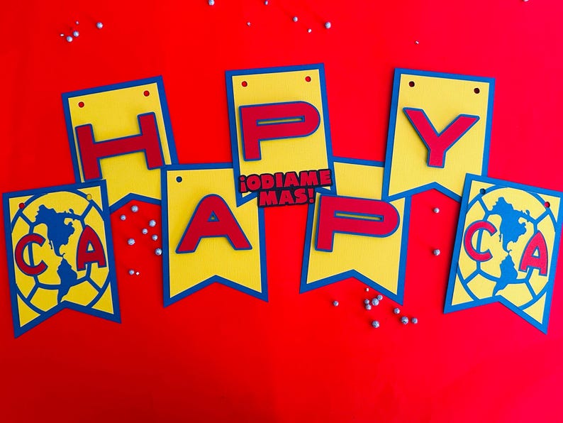 May include: A yellow and blue banner with red letters spelling "Happy" and a soccer ball with the letters "CA" on it. The banner also includes the text "&iexcl;ODIAMOS MAS!" in red letters.