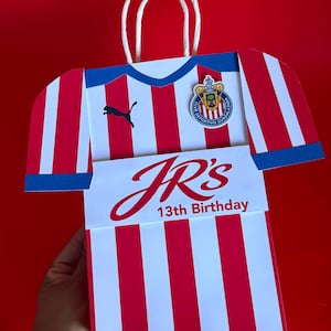 May include: A red and white striped gift bag shaped like a soccer jersey, with blue accents and a white handle. The bag features the Puma logo, a team emblem, and the text "JR's 13th Birthday."