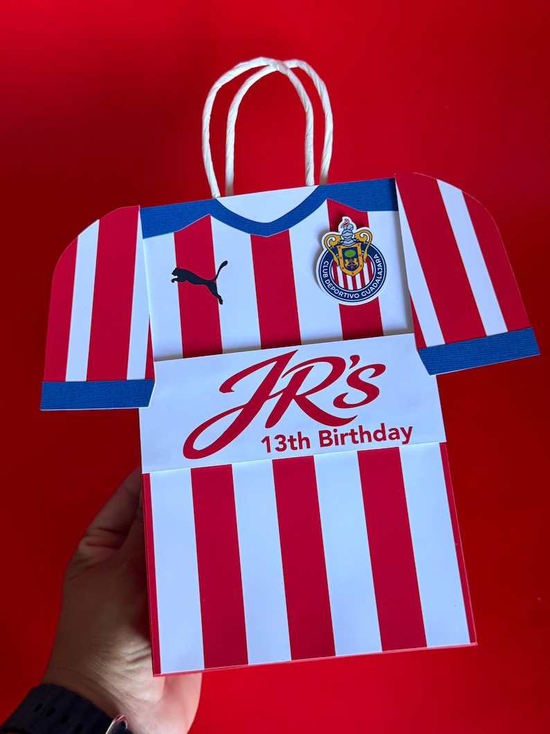 May include: A red and white striped gift bag shaped like a soccer jersey, with blue accents and a white handle. The bag features the Puma logo, a team emblem, and the text "JR's 13th Birthday."