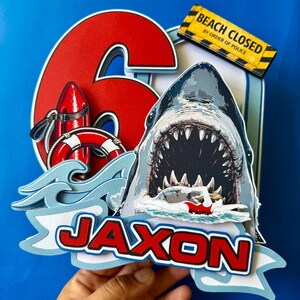 May include: A layered cake topper featuring a large red number 6, a shark with an open mouth, a red life buoy, and a swimmer. A yellow sign reads BEACH CLOSED BY ORDER OF POLICE. The name JAXON is displayed in red letters on a light blue banner.