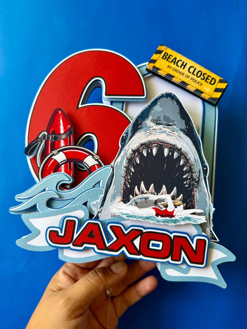 May include: A layered cake topper featuring a large red number 6, a shark with an open mouth, a red life buoy, and a swimmer. A yellow sign reads BEACH CLOSED BY ORDER OF POLICE. The name JAXON is displayed in red letters on a light blue banner.