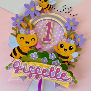 May include: A colorful cake topper featuring cartoon bees, flowers, and the number 1. The topper includes the name "Gisselle" in a decorative font, perfect for a first birthday celebration. The design incorporates yellow, pink, purple, and green.