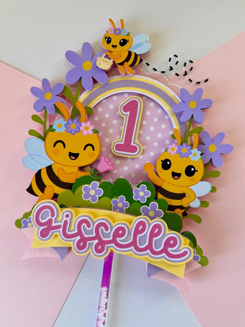 May include: A colorful cake topper featuring cartoon bees, flowers, and the number 1. The topper includes the name "Gisselle" in a decorative font, perfect for a first birthday celebration. The design incorporates yellow, pink, purple, and green.