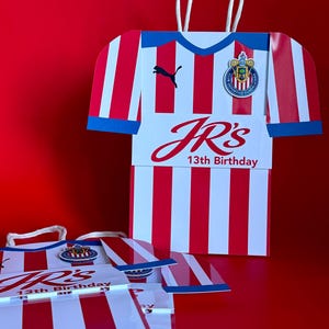 May include: Red and white striped gift bags shaped like a soccer jersey, with blue trim and white handles. The bags feature the text "JR's 13th Birthday" and a team logo. A black Puma logo is also visible.
