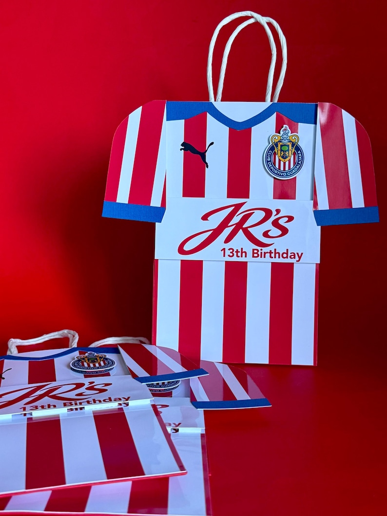 May include: Red and white striped gift bags shaped like a soccer jersey, with blue trim and white handles. The bags feature the text "JR's 13th Birthday" and a team logo. A black Puma logo is also visible.