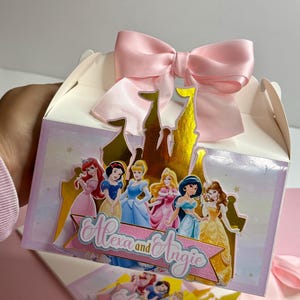 May include: A pink and white favor box with a gold castle design featuring Disney princesses. The box has a pink satin bow and the text "Alexa and Angie" on the front.