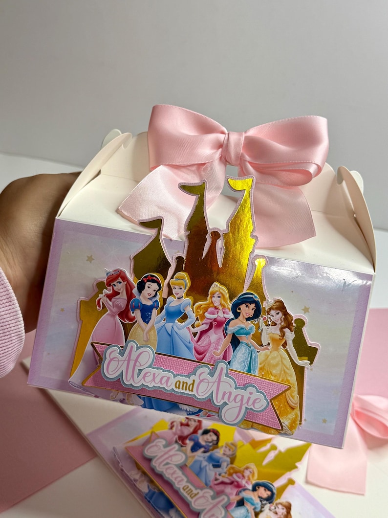 May include: A pink and white favor box with a gold castle design featuring Disney princesses. The box has a pink satin bow and the text "Alexa and Angie" on the front.
