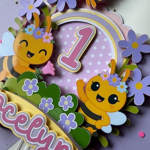 May include: A colorful, layered paper cake topper featuring cartoon bees, flowers, and the number 1. The topper includes the name "Evelyn" in a decorative font, with a pastel color scheme of yellow, purple, and pink.