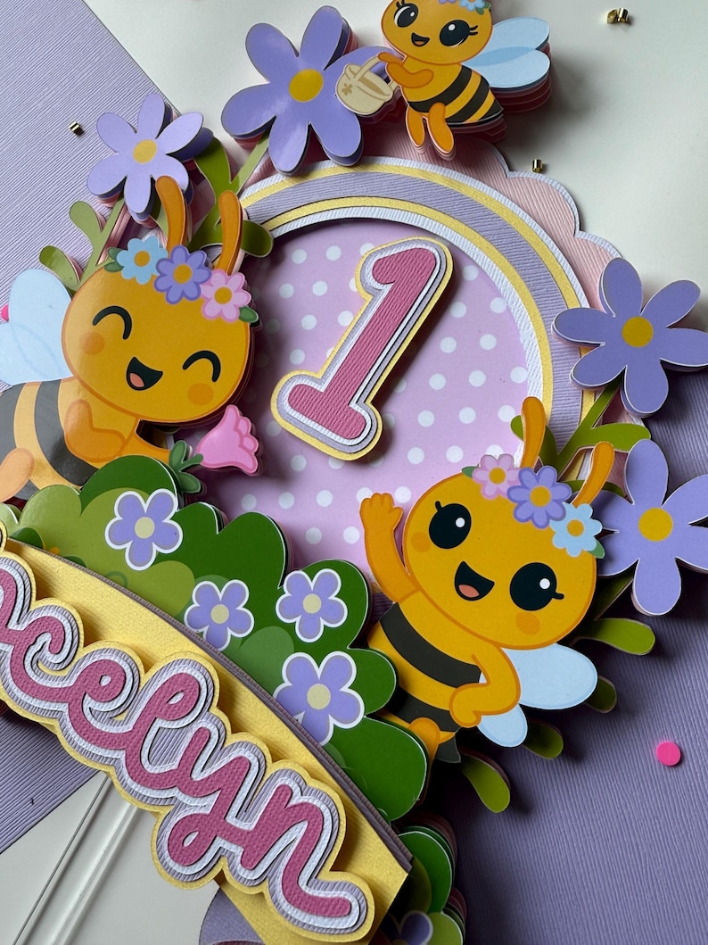 May include: A colorful, layered paper cake topper featuring cartoon bees, flowers, and the number 1. The topper includes the name "Evelyn" in a decorative font, with a pastel color scheme of yellow, purple, and pink.