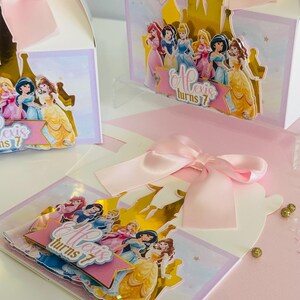 May include: A pink and gold princess-themed party favor box with a pop-up castle design. The box features a gold castle with a pink banner that says "Alexis turns 7". The box is decorated with images of Disney princesses.