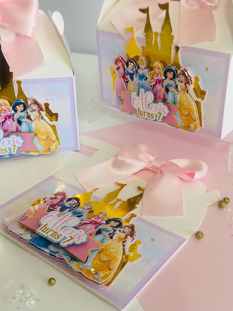 May include: A pink and gold princess-themed party favor box with a pop-up castle design. The box features a gold castle with a pink banner that says "Alexis turns 7". The box is decorated with images of Disney princesses.