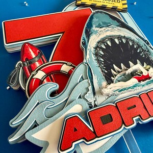 May include: A layered paper cake topper featuring a large red number 7, a shark with an open mouth, a swimmer, a red life buoy, and a yellow sign reading BEACH CLOSED BY ORDER OF POLICE. The name ADRI is partially visible in red block letters.