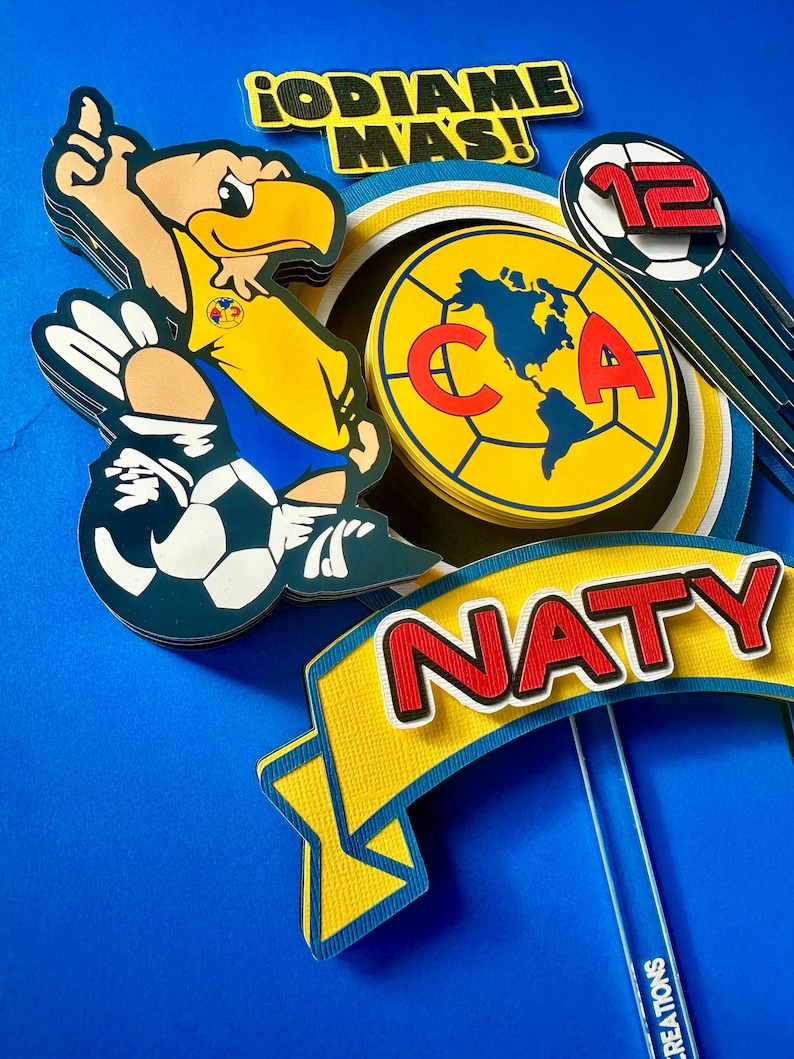 K&ouml;nnte beinhalten: Ein gelber und blauer Tortendekor mit einem Cartoon-Adler, einem Fu&szlig;ball und dem Text "&iexcl;ODIAME MAS!" und "NATY".