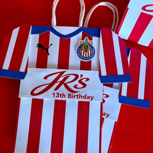 May include: Red and white striped paper gift bag shaped like a soccer jersey, with blue accents and a team emblem. The bag has the text "JR's 13th Birthday" in red. White paper handles are attached.
