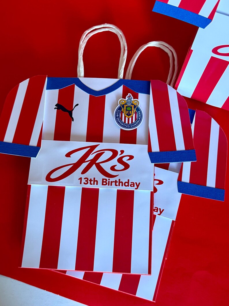May include: Red and white striped paper gift bag shaped like a soccer jersey, with blue accents and a team emblem. The bag has the text "JR's 13th Birthday" in red. White paper handles are attached.